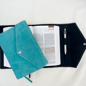 May include: A teal blue leather journal with a black cover and a silver pen. The journal has a flap that closes with a silver button. The cover of the journal has the word "MATTHEW" printed on it.