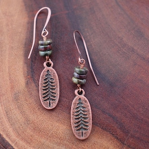May include: Copper earrings with a tree design. Each earring features a copper hook, a cluster of green and brown beads, and an oval pendant with a detailed tree silhouette. The earrings are displayed on a dark wood surface.