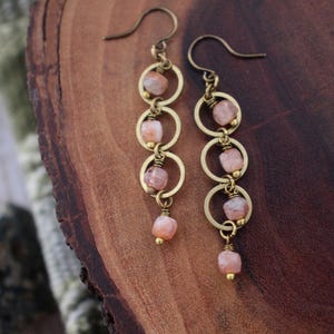 May include: A pair of dangle earrings with a gold-tone finish. Each earring features three connected gold rings, adorned with small, light pink gemstones. The earrings hang from a curved hook. The background is a dark wood surface.