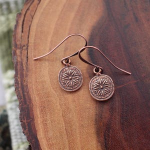 May include: A pair of copper-colored earrings with a compass rose design. The earrings have a hook closure and are displayed on a wooden surface. The earrings are a jewelry item.