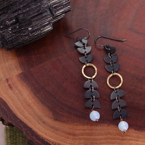 May include: A pair of long, black and gold earrings with a geometric design. Each earring features a series of black half-circles connected by gold rings, ending with a small, white opal bead.
