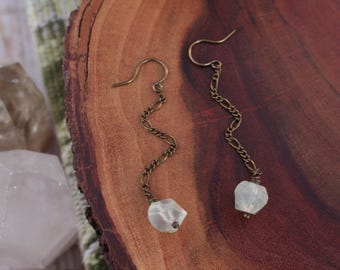 May include: A pair of long, dangling earrings with a white, faceted gemstone at the end of each chain. The chains are made of a brass-colored metal and have a wavy, zig-zag pattern.