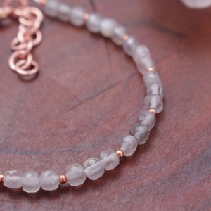 May include: A delicate bracelet featuring faceted, light gray beads interspersed with small copper-colored beads. The bracelet has a copper-colored chain and clasp. The jewelry is displayed on a textured, reddish-brown surface.