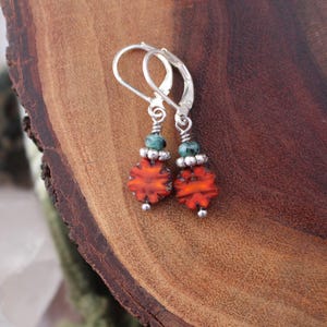 May include: A pair of silver leverback earrings. Each earring features a cluster of small, round silver beads, a turquoise bead, and a vibrant orange, flower-shaped bead. The earrings are displayed on a wooden surface.