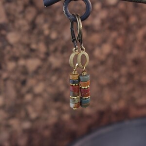 May include: A pair of dangle earrings with a stacked bead design. The earrings feature a mix of earth-toned beads in shades of brown, red, and green, accented with gold-colored metal. The earrings are suspended from a gold-colored hoop.