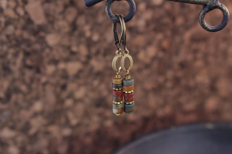 May include: A pair of dangle earrings with a stacked bead design. The earrings feature a mix of earth-toned beads in shades of brown, red, and green, accented with gold-colored metal. The earrings are suspended from a gold-colored hoop.