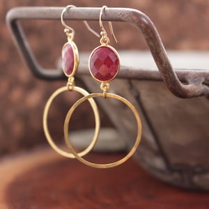 May include: Gold hoop earrings with a dangling red gemstone. The earrings are hanging from a rusty metal hook.