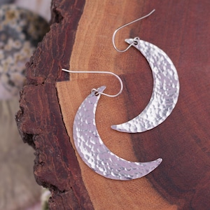 Può includere: Orecchini a forma di mezzaluna color argento con una texture martellata. Gli orecchini sono esposti su una superficie di legno, mostrando il loro design unico. Hanno una chiusura a gancio.