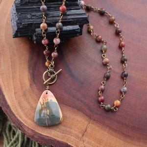 May include: A handcrafted necklace featuring a pendant with a unique pattern of green, pink, and beige. The necklace has a toggle clasp and is strung with small, colorful beads. The chain and clasp are a bronze color.