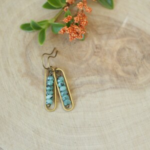 May include: A pair of handcrafted earrings featuring small, light green beads encased in a gold-colored rectangular frame. The earrings hang from a curved hook. The background is a light-colored wooden surface with a sprig of orange flowers.