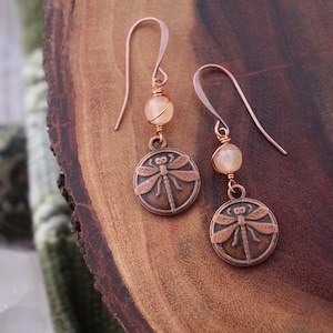 以下が含まれることがあります： 銅色のトンボのイヤリング、ピーチ色のビーズアクセント付き。 イヤリングは、円形のペンダントに詳細なトンボのデザインが特徴です。 イヤーフックはシンプルなフック スタイルです。 木製の表面に表示されています。