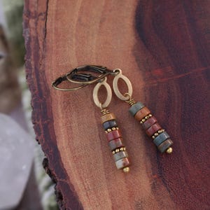 May include: A pair of dangle earrings with a stacked bead design. The earrings feature a mix of colorful, round stone beads in shades of red, brown, blue, and tan. The metal components are gold-toned.