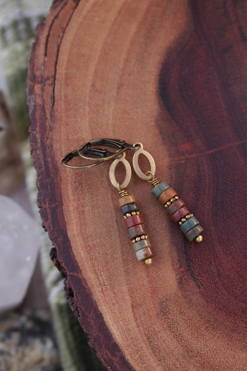 May include: A pair of dangle earrings with a stacked bead design. The earrings feature a mix of colorful, round stone beads in shades of red, brown, blue, and tan. The metal components are gold-toned.
