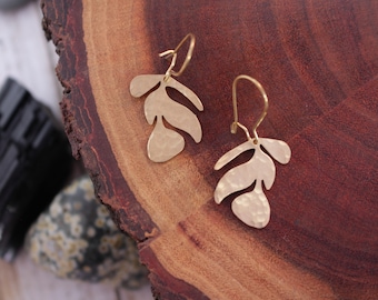 May include: A pair of gold-colored leaf-shaped earrings. Each earring features three textured leaf cutouts suspended from a gold-colored hook. The earrings are displayed on a wooden surface with a dark, natural grain.
