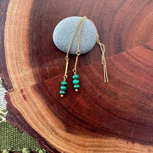 Peut inclure: Une paire de boucles d'oreilles en chaîne dorée avec des perles de pierres précieuses vertes. Les boucles d'oreilles sont présentées sur une surface en bois.