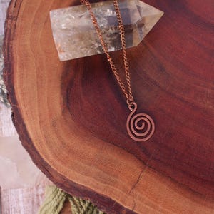 May include: A copper-colored necklace with a spiral pendant rests on a dark wooden surface. The necklace chain is delicate, and the pendant is a simple, elegant design. A clear crystal and other stones are in the background.
