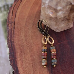 May include: A pair of dangle earrings with a bronze-colored metal design. Each earring features a series of colorful, cylindrical beads in shades of red, brown, and green, accented with small gold-colored beads. The earrings are displayed on a wooden surface.
