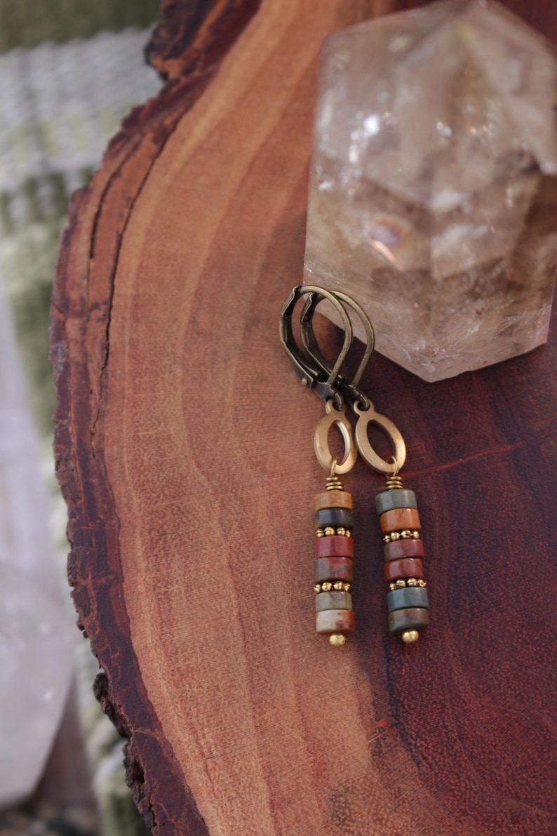 May include: A pair of dangle earrings with a bronze-colored metal design. Each earring features a series of colorful, cylindrical beads in shades of red, brown, and green, accented with small gold-colored beads. The earrings are displayed on a wooden surface.