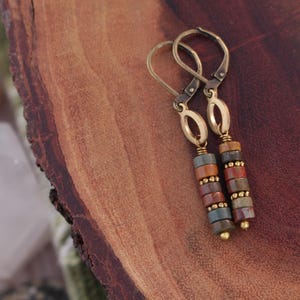 May include: A pair of dangle earrings with a stacked bead design. The earrings feature gold-colored lever-back closures and oval accents. The beads are in shades of red, brown, blue, and gold, creating a colorful and earthy aesthetic.