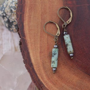 May include: A pair of dangle earrings with a lever back closure. Each earring features a light green, rectangular bead with dark green marbling, accented with small, dark grey beads. The earrings have an antique brass finish.