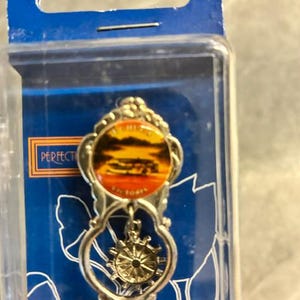 May include: A silver-toned souvenir spoon featuring a circular emblem with a sunset landscape and the text "ECHUCA VICTORIA". Below the emblem, a small silver ship's wheel charm hangs within a decorative openwork handle design, all encased in plastic.