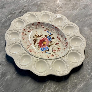 May include: A white ceramic egg serving platter with a scalloped edge and twelve circular indentations. The central oval area features a hand-painted floral design with a large pink flower, blue blossoms, and green leaves, surrounded by red scrollwork.