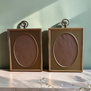 May include: Two rectangular brass picture frames with rounded corners and an oval-shaped inner display area. Each frame features a circular hanging ring at the top centre, finished in a polished metallic gold tone with a smooth, reflective surface.