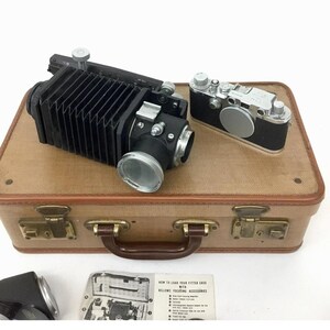 May include: A vintage brown leather suitcase with two silver latches and a handle. The suitcase is open and contains a black bellows camera and a silver rangefinder camera. The suitcase also includes a small instruction booklet titled "How to Load Your Fitted Case with Bellows Focusing Accessories".
