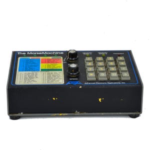May include: A black "The MorseMachine" device with a blue and white panel, knobs, and a keypad. The panel displays text and color-coded sections. The keypad has number and letter keys. The device is labeled "Advanced Electronic Applications, Inc."