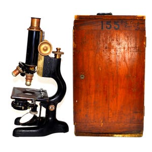 May include: An antique microscope with a black and brass finish, alongside a wooden case. The microscope features multiple lenses and adjustment knobs. The rectangular wooden case has the number "155" printed on it.