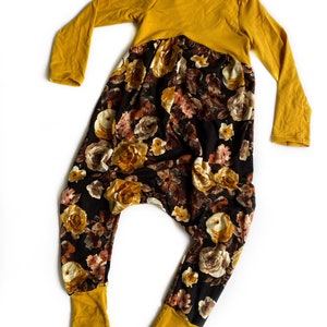 May include: A mustard yellow and black floral print jumpsuit with long sleeves and a gathered waist. The jumpsuit has mustard yellow cuffs and a mustard yellow top.