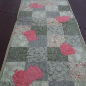 May include: A quilted table runner featuring a patchwork design with squares in shades of green, gray, and white. The runner is adorned with appliqued red apple shapes and has a green border. The runner is approximately 12 inches wide and 48 inches long.