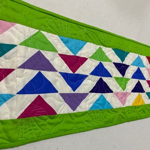 May include: A colorful table runner with a green border and a pattern of triangles in various colors, including pink, blue, purple, red, yellow, and green.