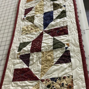 May include: A white quilted table runner with a red border. The runner is decorated with a repeating pattern of colorful fabric squares in shades of red, green, blue, and yellow. The fabric squares are arranged in a geometric pattern.