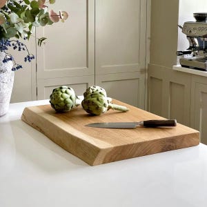 May include: A light brown rectangular wooden chopping board with a natural edge, two artichokes, and a stainless steel knife with a dark brown handle. The board is on a white surface, with a kitchen setting in the background.