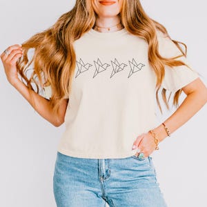 May include: A cream colored t-shirt with a graphic of five origami birds in a row. The birds are black and outlined.