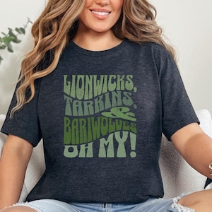 May include: Dark grey t-shirt featuring the text "Lionwicks, Tarkins, Bariwolves, OH MY!" in green hues. The design has a retro, groovy style, with a stylized illustration of a creature.