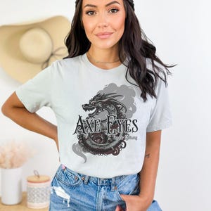 May include: A light gray t-shirt with a black and gray dragon graphic and the text "Axe Eyes" and "Beyond Saluting".