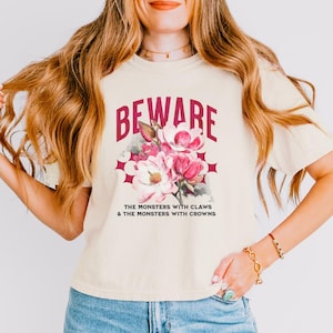 May include: A cream-coloured t-shirt featuring the maroon word "BEWARE" above a floral design. The design includes pink and white flowers with the text "THE MONSTERS WITH CLAWS & THE MONSTERS WITH CROWNS" below the flowers.