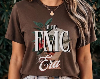 May include: Brown t-shirt with a graphic design featuring a woman's face with a flower covering one eye. The text "in my FMC Era" is printed on the shirt.