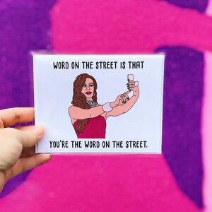 May include: A white card with a pink and white illustration of a woman taking a selfie. The text on the card reads "Word on the street is that you're the word on the street."