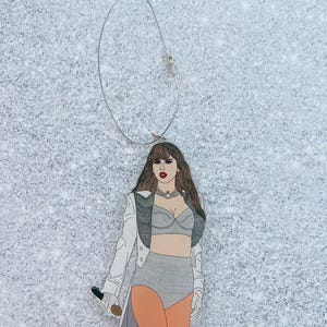 May include: A white and silver Christmas ornament featuring a cartoon illustration of a woman wearing a white jacket, a silver bra, and a silver skirt. She is holding a microphone and has long brown hair.