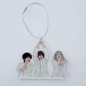 Puede incluir: Adorno con una ilustración estilizada de tres personas con trajes blancos. Las figuras tienen diferentes peinados y poses, con una cuerda plateada para colgar. El fondo es blanco.