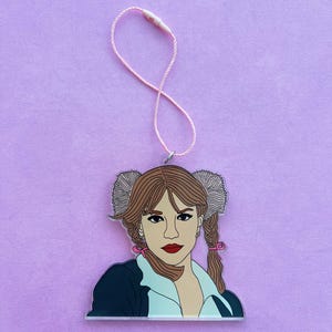 May include: An illustrated character with brown hair, wearing a blue jacket and white shirt, with white fluffy earmuffs and pink bows in her braids. The character is hanging from a pink string.