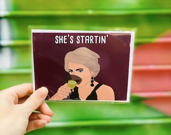 Puede incluir: Una tarjeta de felicitación con una ilustración de dibujos animados de una mujer bebiendo de una copa de martini. La tarjeta tiene un fondo burdeos y el texto "SHE'S STARTIN'" en blanco.