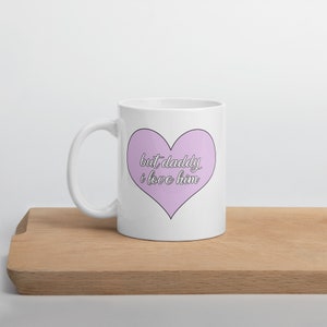 May include: White ceramic mug with a pink heart design. The heart has the text "but daddy i love him" in black.