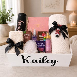 May include: A white wooden gift crate labeled Kailey containing a black tumbler, a pink 2026 planner, two cream-colored textured blankets tied with black ribbons, a floral hand cream, a Kleenex pack, Purell, M&Ms, Body Refresh wipes, and a body mist.