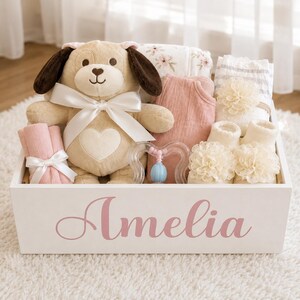 Peut inclure: Une bo&icirc;te cadeau en bois blanc avec le pr&eacute;nom Amelia imprim&eacute; en &eacute;criture rose. &Agrave; l'int&eacute;rieur se trouvent un chien en peluche beige avec un ruban blanc, un ensemble en tissu rose, une couverture &agrave; fleurs, un hochet en plastique et deux chaussons.