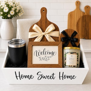 May include: A white wooden crate labeled Home Sweet Home containing a black tumbler, a striped kitchen towel, a wooden cutting board with a cream ribbon, a framed Welcome sign, and a Tobacco & Rose candle with a black ribbon on its lid.