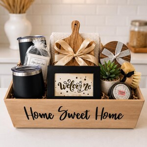 May include: A wooden gift crate labeled Home Sweet Home containing two black tumblers, a wooden cutting board with a gold ribbon, a black picture frame with the word Welcome, a small potted succulent, a round wooden coaster, and a scrub brush.